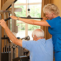 A senior man uses a machine to exercise in a rehab center gym with a physical therapist helping him